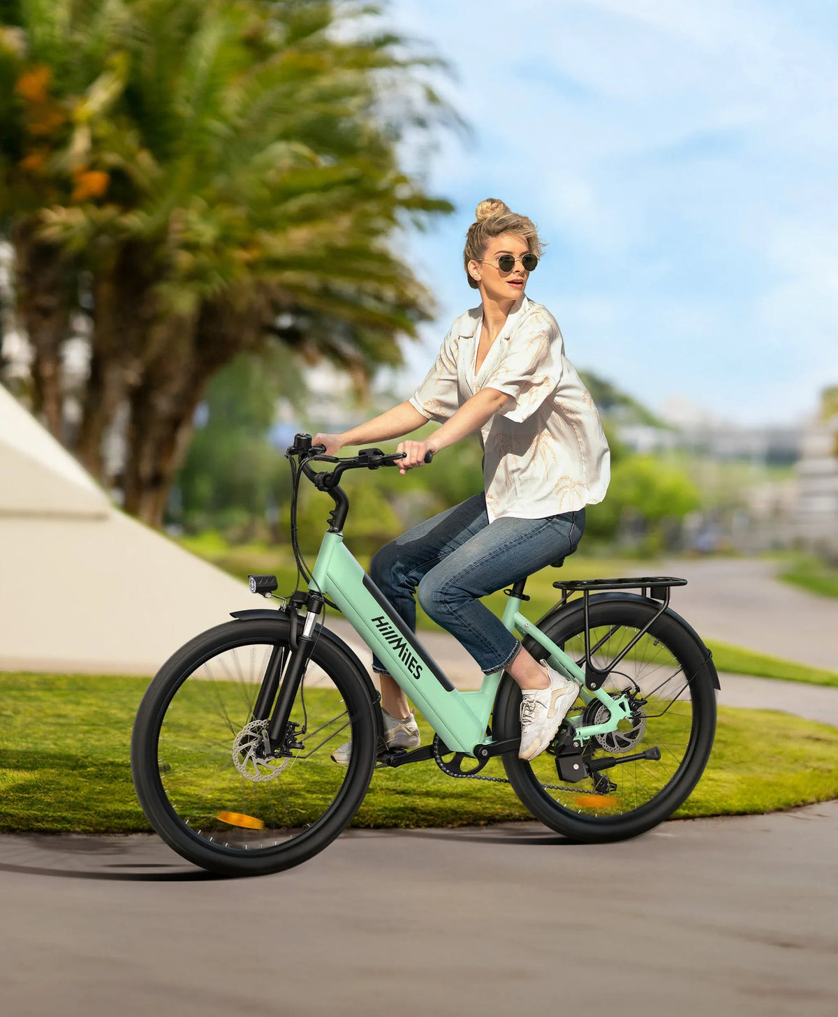 Vélo de ville HillMiles vert menthe, avec porte-bagages, conduit par une femme sur une route bordée de palmiers